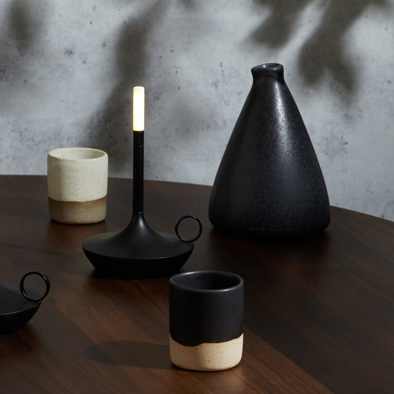 Two black Wick LED tipped candleholder shaped portable lights set on top of a round wood table with two small ceramic cups and a taller black vessel to the left.