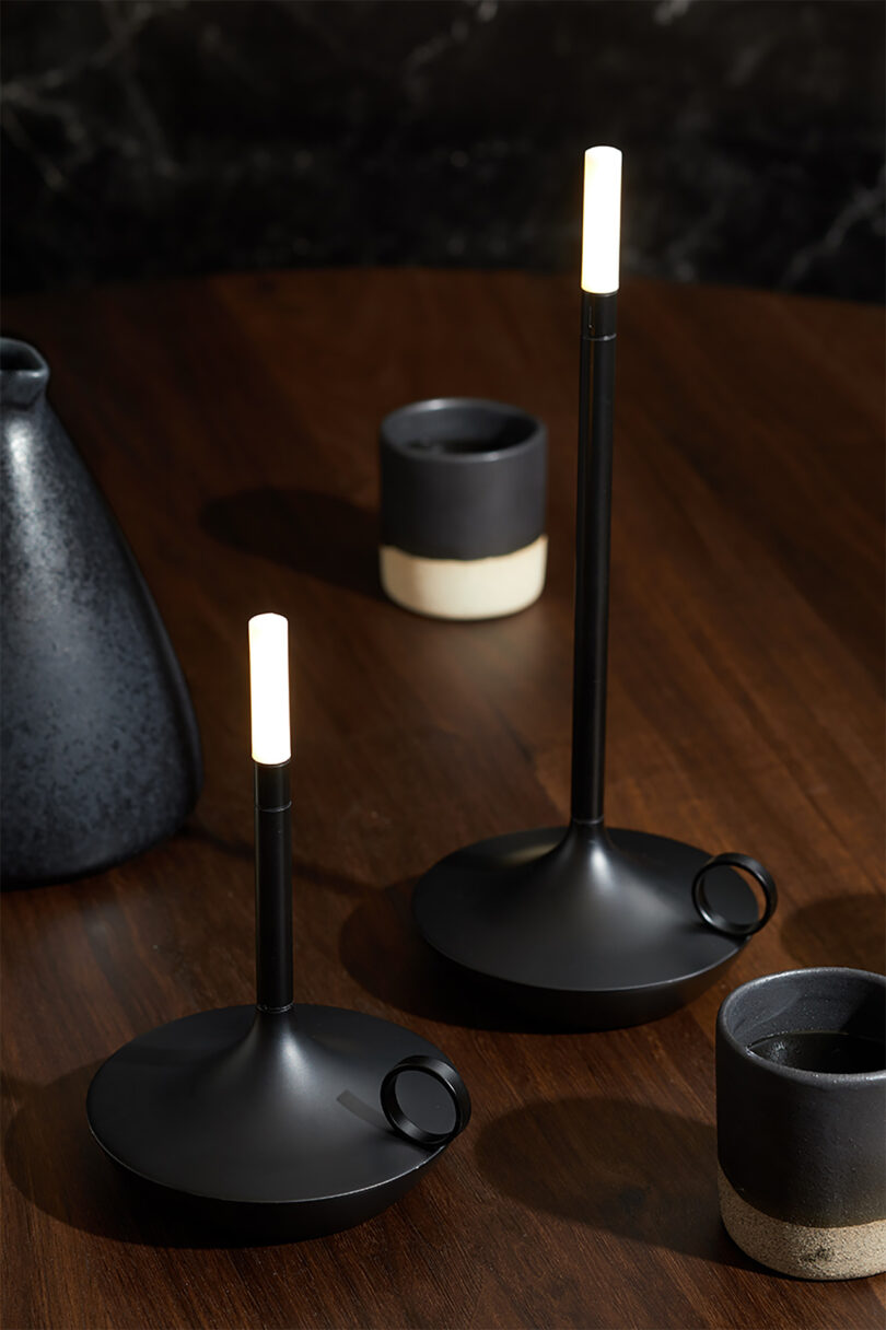Two black Wick LED tipped candleholder shaped portable lights set on top of a wood table with two small ceramic cups and a taller black vessel to the left.