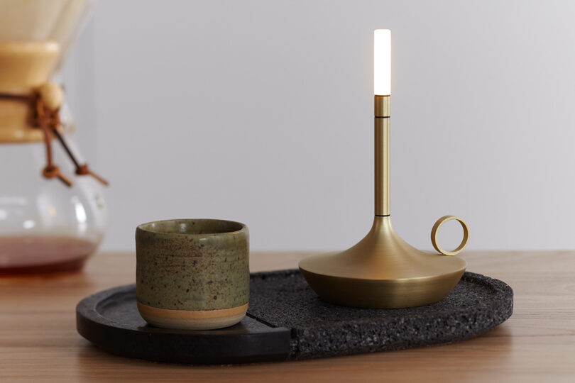 LED tipped Wick candleholder shaped portable light set on top of a oval stone platter next to a small ceramic green coffee cup with Chemex coffee vessel in the background.