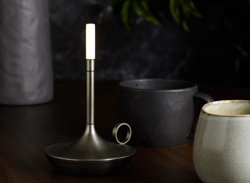 One graphite Wick LED tipped candleholder shaped portable light with one ceramic cup and a dark coffee or tea mug to the right.