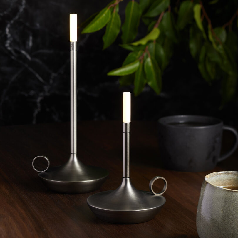 Two graphite Wick LED tipped candleholder shaped portable lights set on top of a wood table with two small ceramic cups and a houseplant in the background.