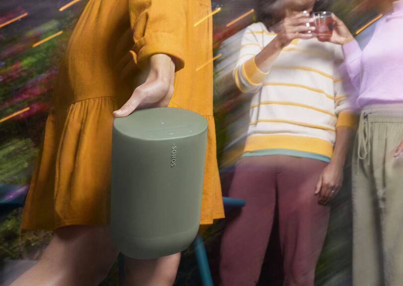 Woman in yellow smock dress carrying Olive Sonos Move 2 wireless speaker at an outdoor backyard shindig with two people on her right toasting one another with plastic drink cups.