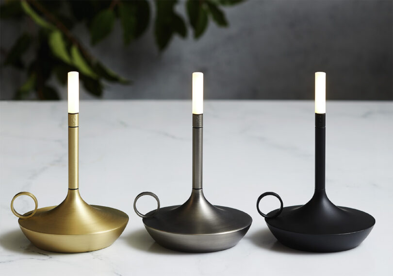 All three colors of the Wick S echargeable light in brass, silver and black. Shaped like a candle holder and tipped with a white 1 watt LED with a circular finger holder.