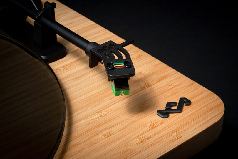 Cropped close up of House of Marley's Stir it Up Lux Bluetooth Turntable with bamboo plinth and dark glass platter and Jamaican color tipped record needle cartridge and tone arm against an all-black background.