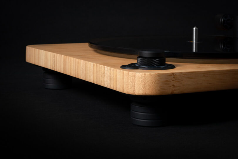 Cropped close up of House of Marley's Stir it Up Lux Bluetooth Turntable's knurled dial controls, with bamboo plinth and dark glass platter and Jamaican color tipped record needle cartridge and tone arm against an all-black background.