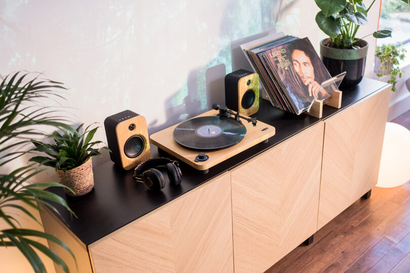House of Marley's Stir it Up Lux Bluetooth Turntable with two bookshelf speakers, headphone, and vinyl record display set across black top console with herringbone wood doors, with several potted houseplants surrounding the stereo setup.