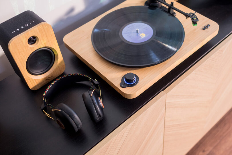 House of Marley's Stir it Up Lux Bluetooth Turntable with two bookshelf speakers, headphone, and vinyl record display set across black top console with herringbone wood doors. Record is on turntable with tone arm in "off" setting.