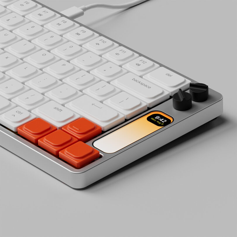 Angled view of four orange arrow keys with two dial controls in black in the upper right hand corners with the keyboard's small LCD screen displaying the time of 9:42. Keyboard is in plugged in USB-C connection mode with a cable snaking from the back.