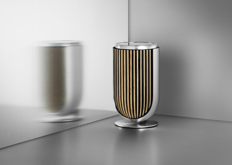 Aluminum and wood slat covered Beolab 8 speaker set upon the floor near a reflective aluminum wall surface. Reflection is blurry.