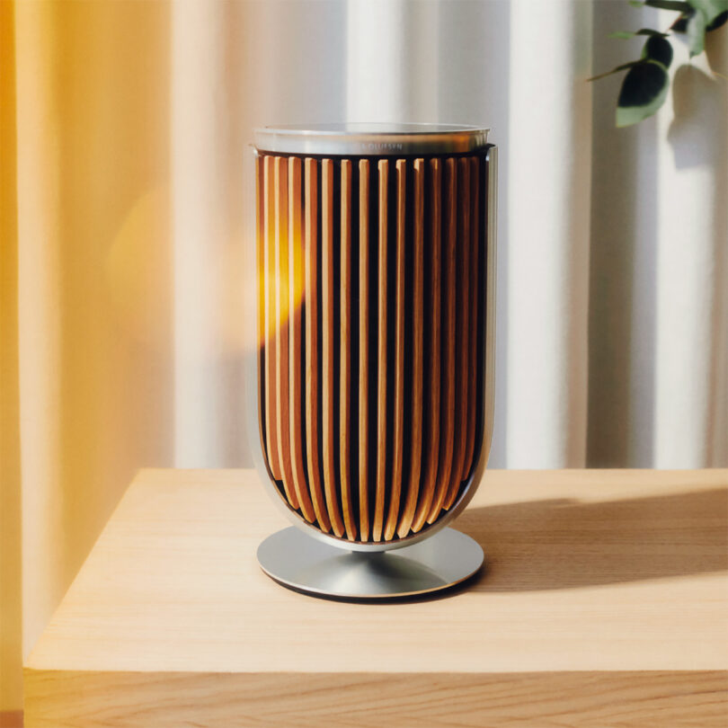 Single Beolab 8 speakers with wood slat covers set on wood console surface with the hint of a houseplant and curtains in the background. Photo has light leaks of orange light coming from the left.