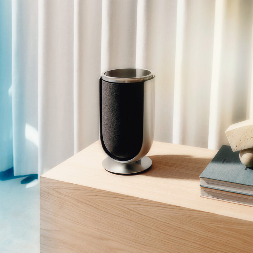 Single Beolab 8 speaker with fabric cloth cover in black set across wood console. Two hardcover books are stacked to the right with a small sculptural paperweight on top, and white curtains in the background.