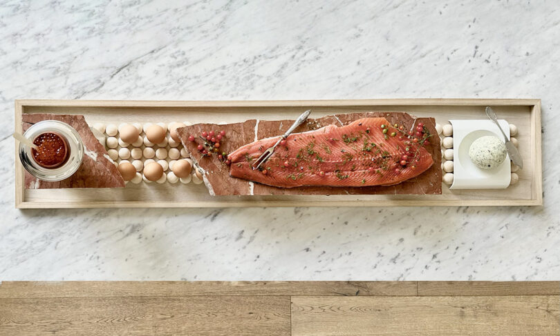 Long board with homemade gravlax salmon displayed across with ball of herbed butter to right and bowl of salmon roe to garnish on the left. Set across a marble countertop.