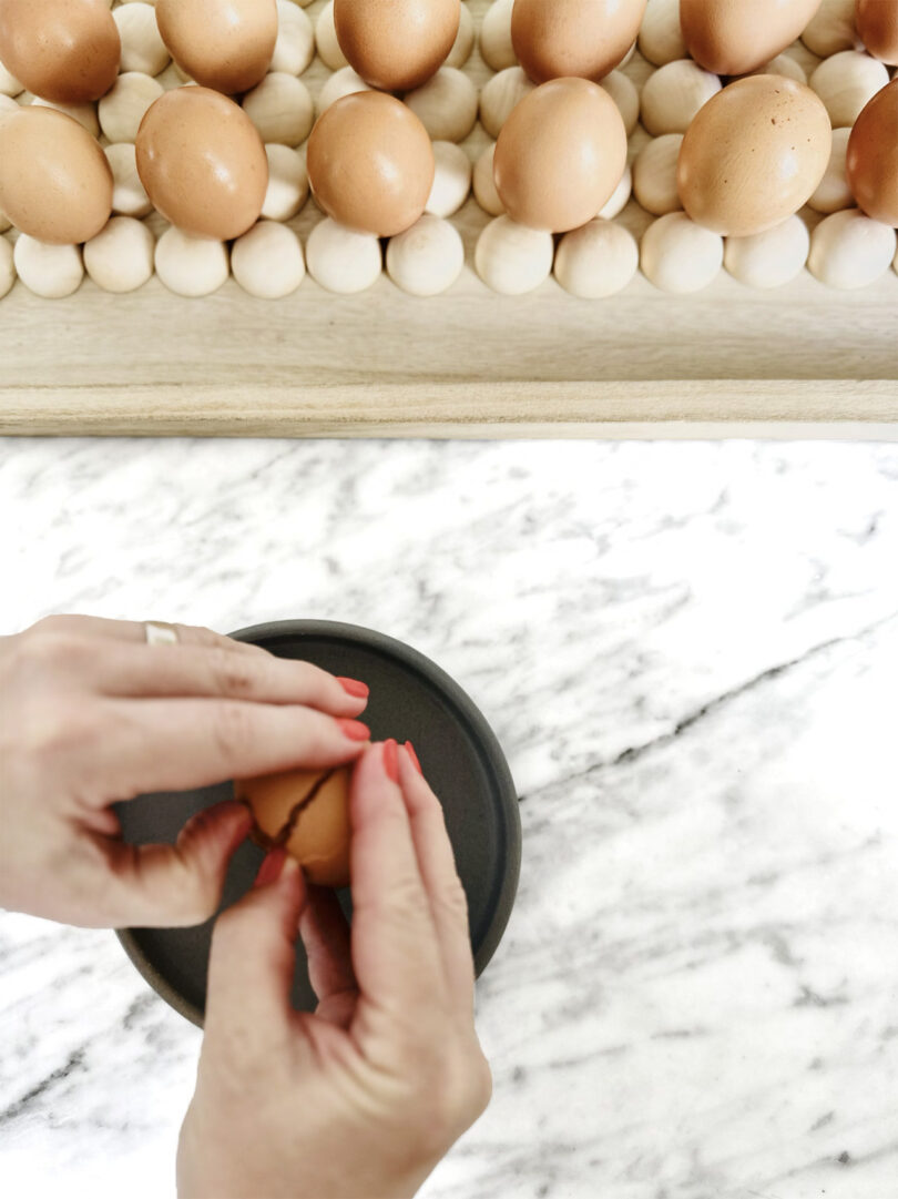Overhead shot of person's two hands cracking open an egg over marble countertop with 10 additional eggs visible at top of photo.