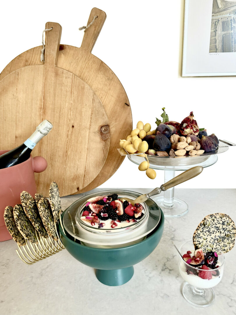 Bowl of homemade yogurt garnished with berries and figs with toil crackers to the left and whole nuts, figs and open pomegranate to the right. In the background are two round handled cutting boards.