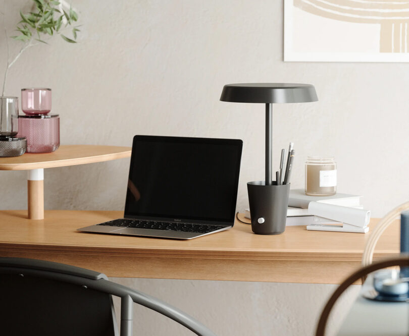 Black Umbra CUP desk lamp with pens and pencils stored in its cup-styled base across a wood desk with open MacBook Air to its left. Additional home accessories are staged across the desk, including a white stapler, canldle, books, and some tinted glassware.