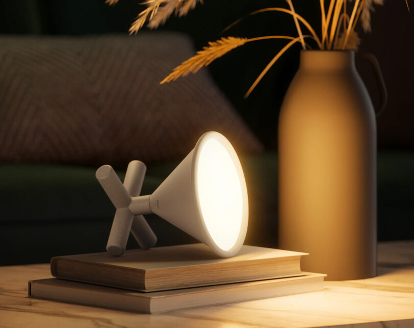 Gray Umbra CONO LED light set on its side on top of two books, glowing warm white light across the books and a nearby vase filled with an arrangement of ornamental grass stalks.