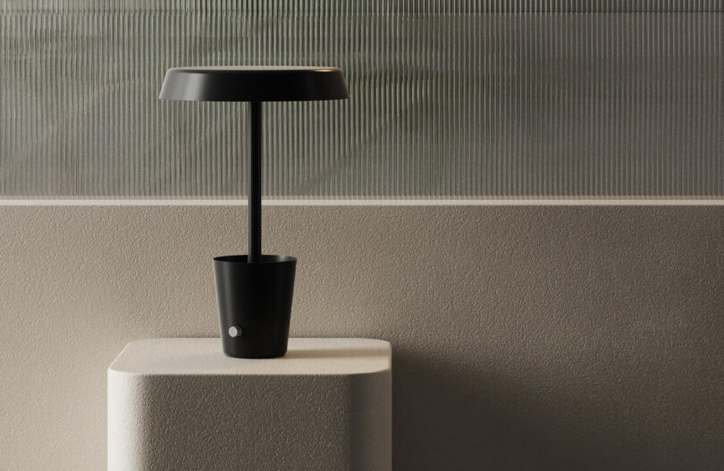 Black CUP LED lamp set on a stone-like warm tan pedestal. Lamp's cup-shaped based is empty, with its dial control facing toward the left at an angle.