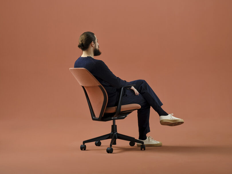 Bearded man with man bun in dark blue clothing seated in the Vitra ACX task chair against a salmon-orange hued background. He is turned facing away from viewed at a slight angle with legs crossed.