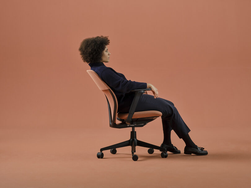 Woman with curly dark hair in dark blue clothing leaning back in the Vitra ACX task chair against a salmon-orange hued background.