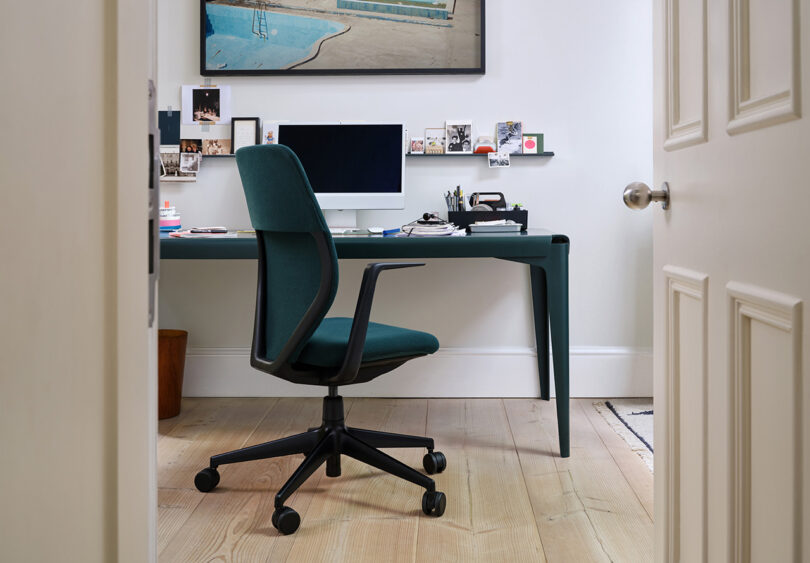 Viewing into a home office with the door ajar of a dark green and black Vitra ACX office chair behind dark green desk with Apple iMac and floating wall shelf decorated with photos and other miscellaneous items.
