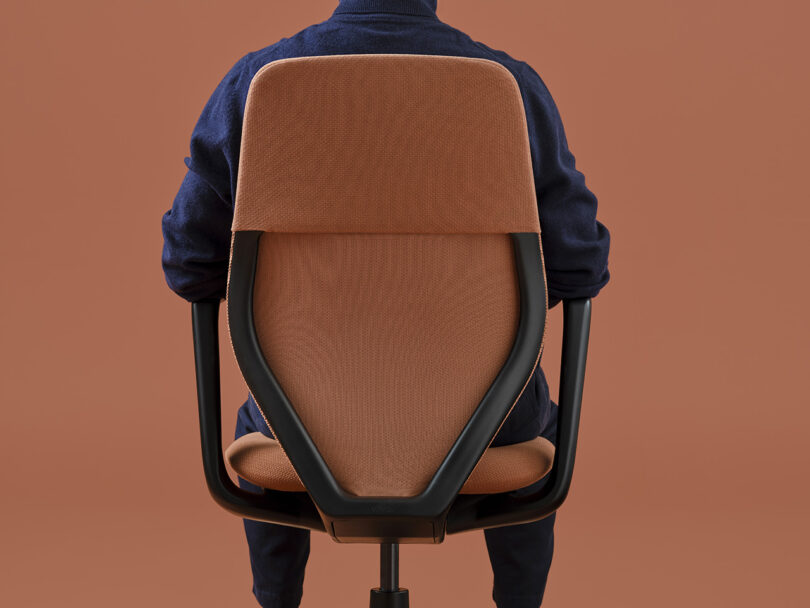 Person's back torso in dark blue clothing seated in the Vitra ACX task chair against a salmon-orange hued background.