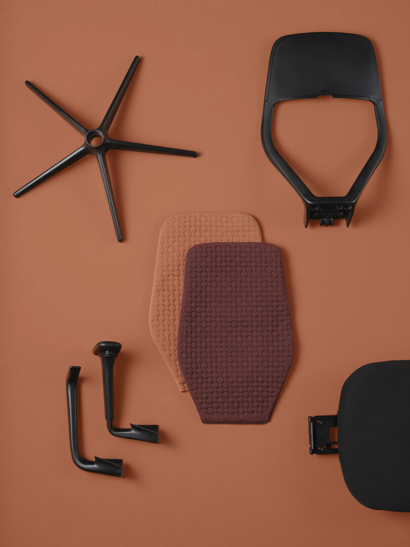 A disassembled Vitra ACX task chair against a salmon-orange hued background with two different knit fabric cover back options shown in dark raspberry hue and another in salmon.