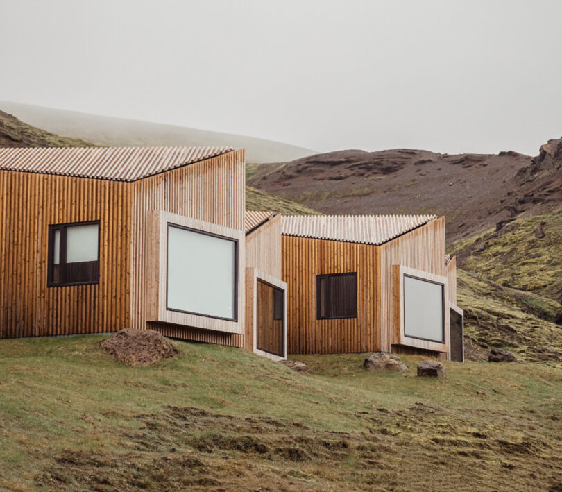 Two of the larger wood clad suites set amongst the rugged Icelandic mountain vistas, each featuring large windows facing the grass covered landscape, and angled rooflines.