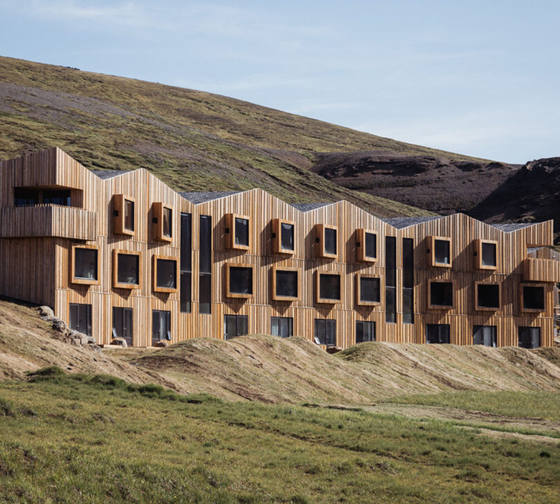 View of the Highland Base - Kerlingarfjöll wood clad hotel exterior, with each room featuring an angled windows looking out onto the Icelandic summer landscape with grasses growing along mounds.