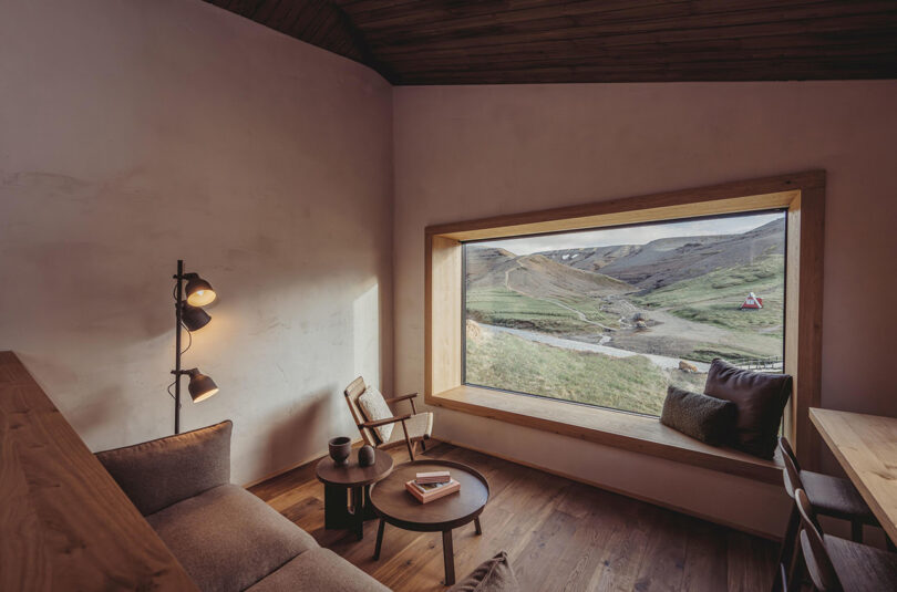 Warm interior shot of one of the Highland Base - Kerlingarfjöll's suites furnished with modern Scandinavian designs. A large window affords a wide view of the Icelandic landscape of rolling hills covered in summertime grass with a cool gray light.