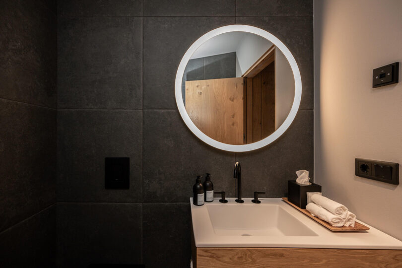 Contemporary design bathroom with dark stone tile walls, small sink with rolled hand towels, toiletries, and a small circular wall mirror over the sink.