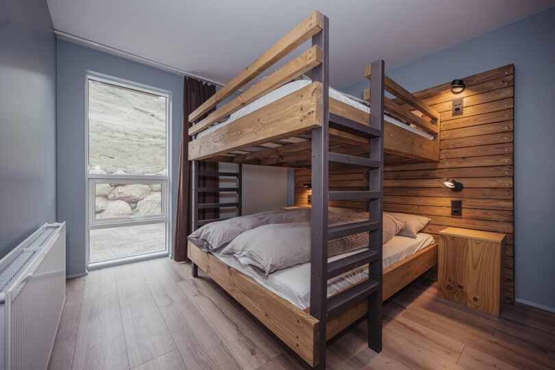 Photo of interior hostel room for families furnished with a queen size bunk bed with horizontal wood paneling along the length of the bed's adjoining wall, and a narrow-tall window looking outside parallel to the bed. Across from bed is a long furnace heater powered by geothermal energy.