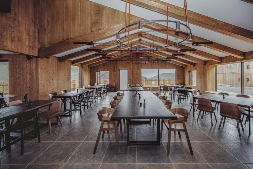 Interior shot of a cavernous 80-seat Highland Base Restaurant and Lounge, with long dining table seating up to 8 people in the center with long overhead lights stretching above, stone tile flooring and wood clad interior walls.