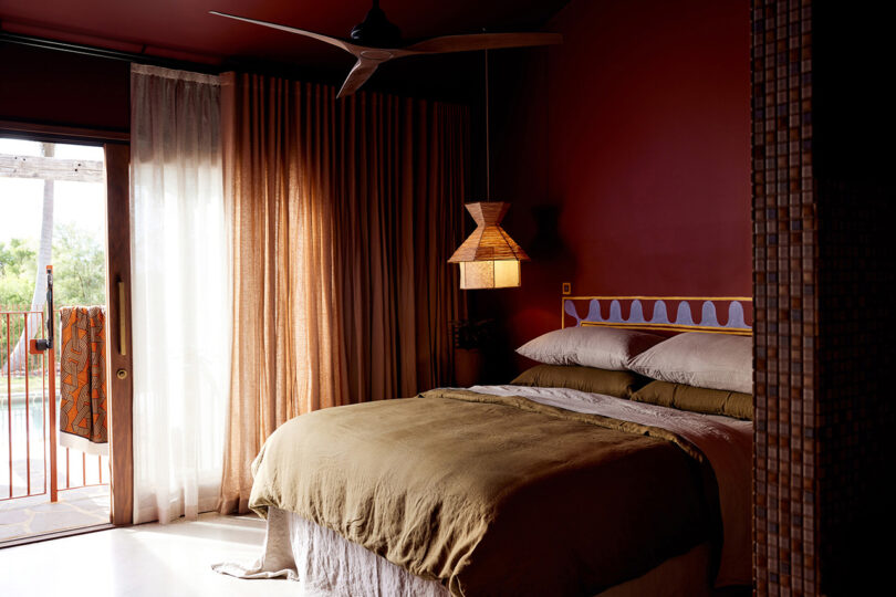Dark red/magenta bedroom with small beside pendant lamp hanging in the left side corner near king side bed with stacked pillows and olive duvet, with drapery covered windows looking out to a pool.