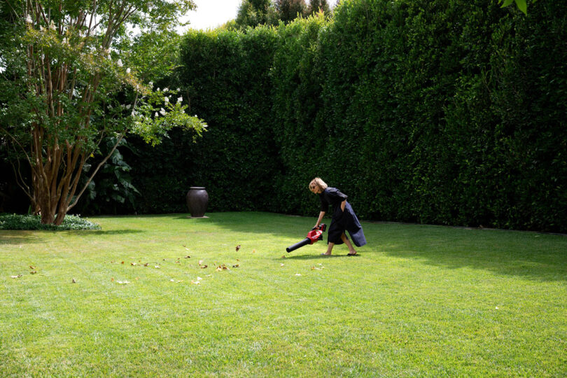 a woman wearing a black dress uses a leaf blower on a pristine green lawn