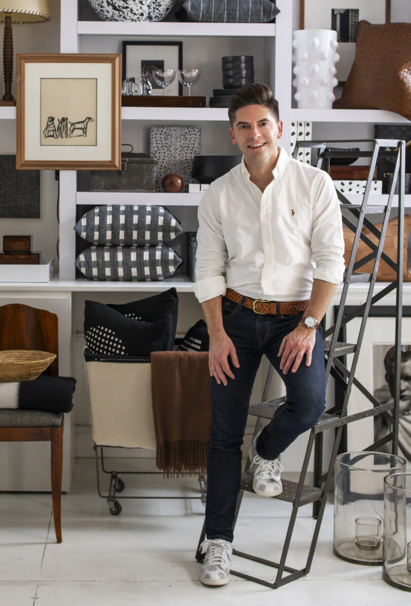 light-skinned man poses in front of a large shelving unit filled with homewares