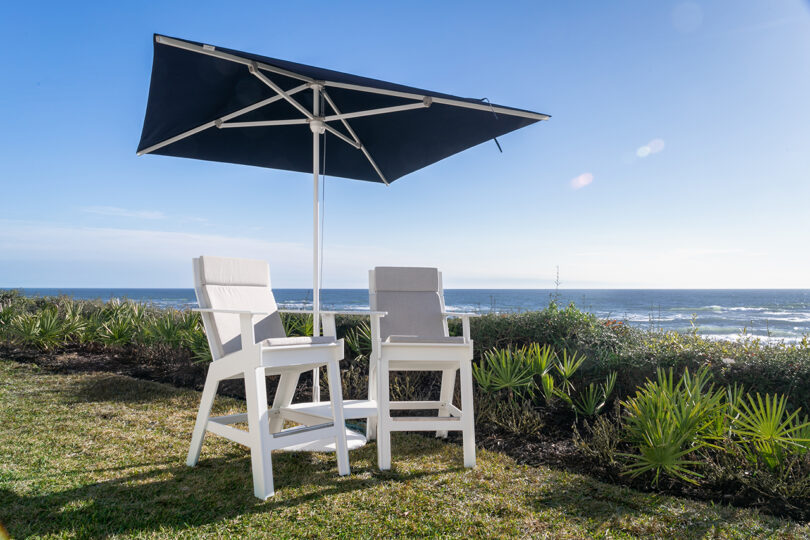 two white hi-rise chairs under a square umbrella overlooking the water