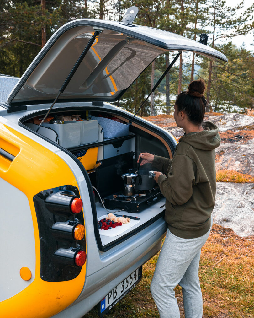 angled back view of yellow and white camper with backend opened with woman cooking in built-in kitchen