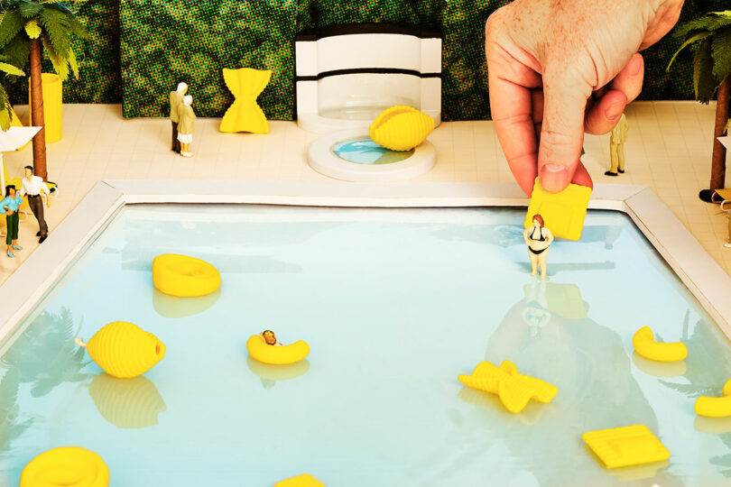 miniature scene of swimming pool with tiny pasta-shaped floats with random people and a real hand poking in holding a piece