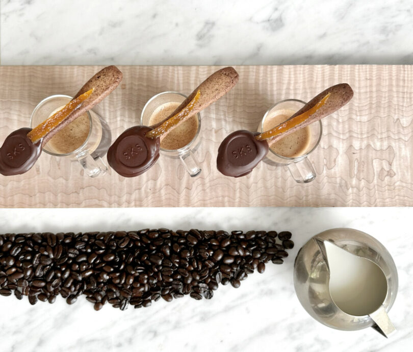 Spoon-shaped cookies with chocolate covered ends with SKS monogrammed into chocolate set across their own small cups of espresso. Espresso coffee beans are gathered along the bottom edge with small metal creamer pourer to the right.