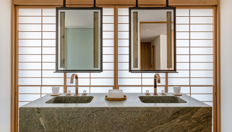 A single green marble covered bathroom counter with twin sinks, faucets and mirrors. A shoji screen illuminates the background with soft white light.
