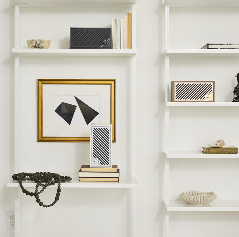 Three Tivoli Audio Modern Two Digital audio speakers staged on a modern white bookshelf. One in black sits on top holding up five small hardback books, the second is positioned vertically on top of four books with a small piece of framed abstract black and white artwork behind it, and a third wood and brass speaker sits to the right horizontally.