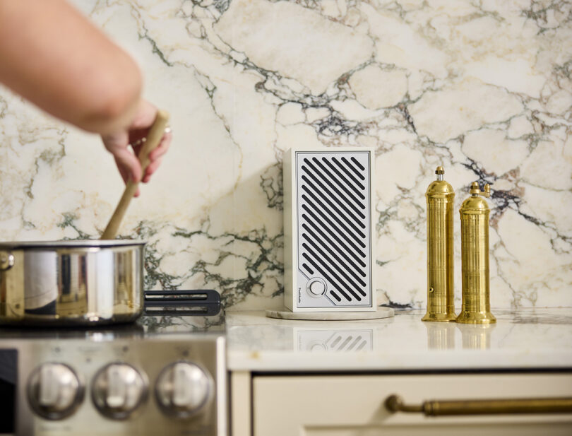 White and silver Tivoli Audio Modern Two Digital staged in kitchen to the left of someone stirring a pot of food set on a range. A set of tall pepper and salt grinders sit to the right of the speaker, with a veined marble backsplash behind it.