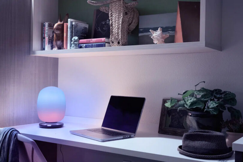 Smaller SKYVIEW 2 table lamp set across white desk set near open MacBook with floating shelf overhead with books and small decorative items. Lamp is glowing in a pink to blue gradient.