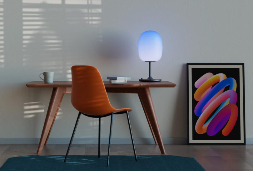 SKYVIEW 2 PRO lamp set on midcentury wooden desk with splayed legs and Eames-inspired shell back chair in orange. The lamp is glowing a gradient of white and blue, with a nearby framed Apple-inspired graphic poster print on the floor to the right of the desk. Against the walls are the shadows of shutter blinds.