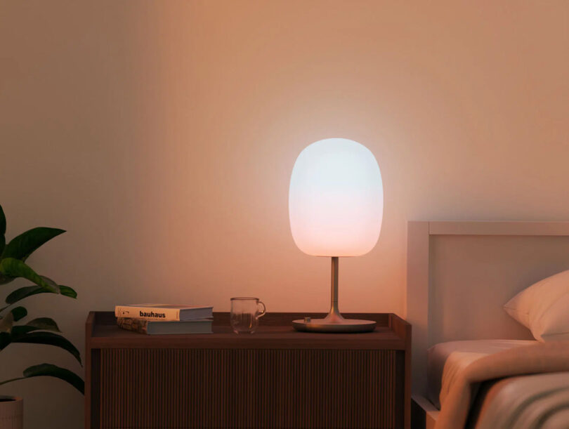 Bedside lamp on aluminum base pedestal with glass globe diffuser, glowing a gentle white and pink light washing across the bedroom's walls, nearby beds and beside dressed with books and small glass across the top.