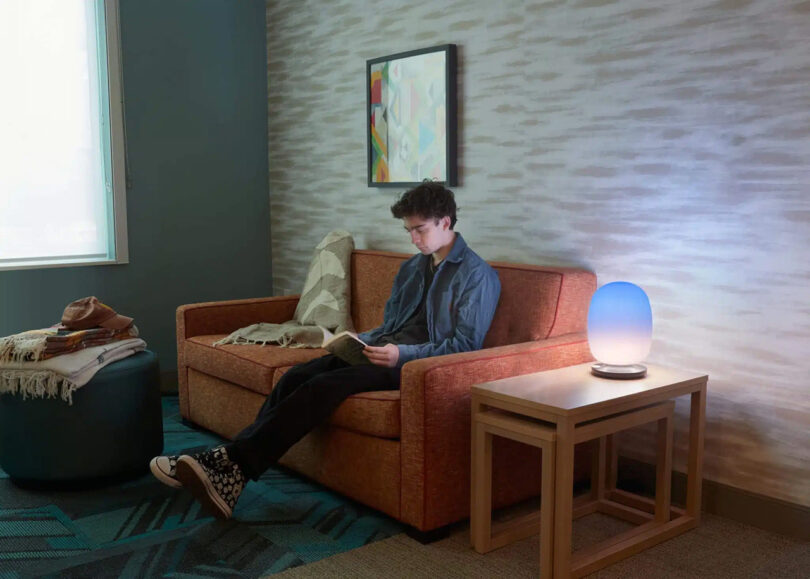 Young man in blue long sleeved top, black jeans and sneakers reading a book on a red loveseat, with a small set of nesting side tables topped with SKYVIEW 2 lamp glowing from the corner.