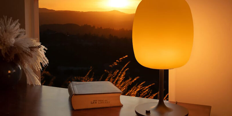 A warm orange sunset seen outside a window is mimicked in hue by the SKYVIEW 2 Pro table lamp. A book stamped with "Law Library" is stamped across the bottom pages.