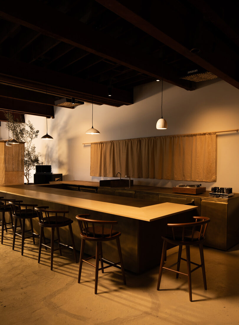 Front angled view of the entire Blue Bottle Studio wood and brass bar with a coffee dyed fabric backdrop and8 walnut seating, illuminated with overhead pendant lighting.