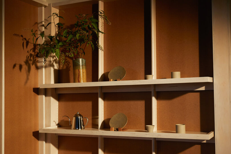 Shallow wall mounted bookshelf displaying Japanese ceramics, coffee pour-over kettle, and ceramic vase with a cut branch of possible coffee leaves and berries. Walls are coffee stained in color.