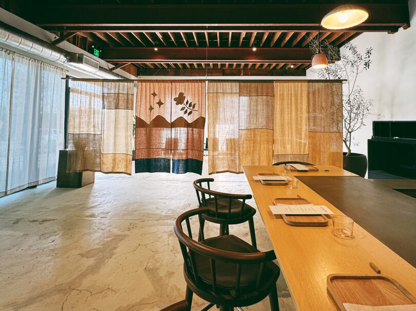 View from back of Blue Bottle Studio showing the wall length expanse of coffee dyed fabric partition dividing the front entrance and the main seating/serving area with long wood and brass surface bar, with walnut seats for guests.